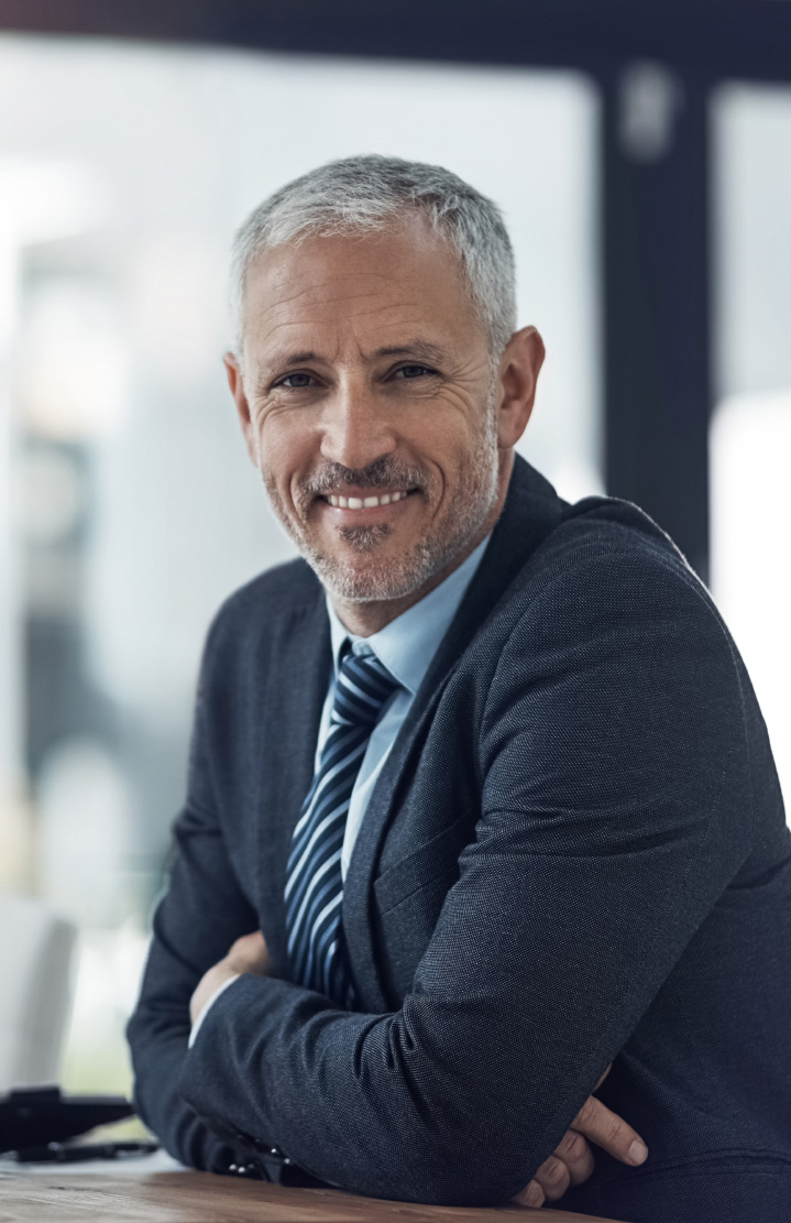 Grey-haired man in a dark suit and striped tie smiling, arms folded on a table