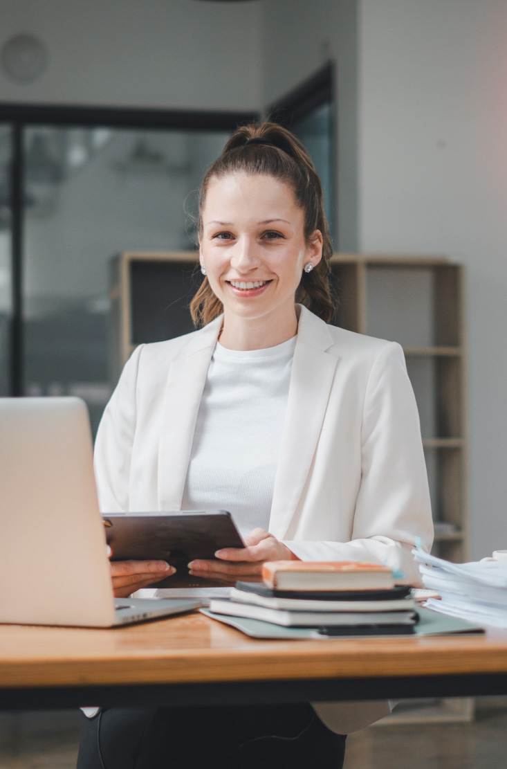 Woman in a white blazer smiling at a desk with a tablet, laptop and stacked books