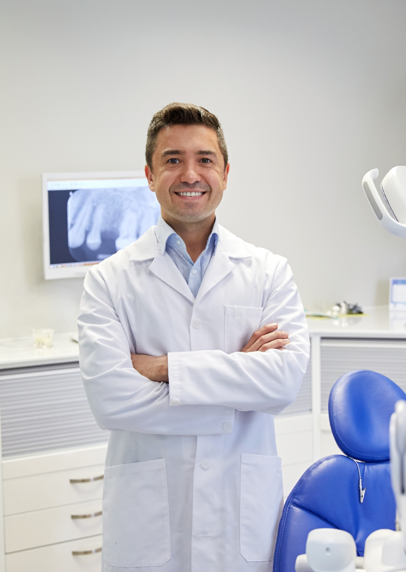 Dentist in a white coat with arms crossed beside a blue treatment chair and dental X-ray screen