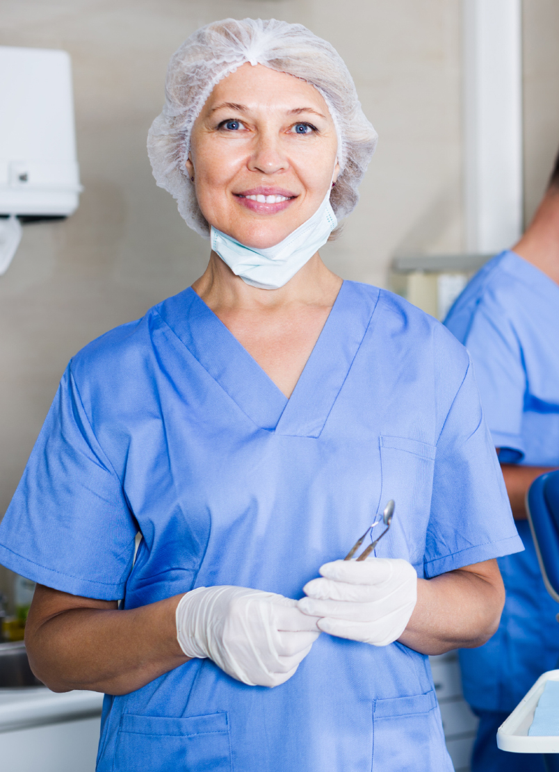Smiling clinician in blue scrubs, hair cap and gloves holding a dental instrument, colleague working behind her