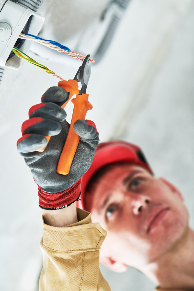 Electrician in a red hard hat and gloves using pliers on wires at a ceiling junction box