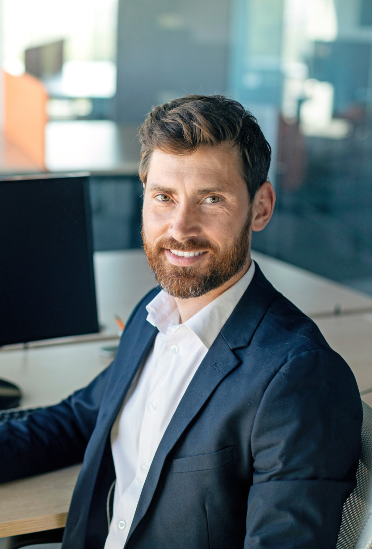 Bearded man in a navy suit jacket smiling at his desk beside a computer monitor