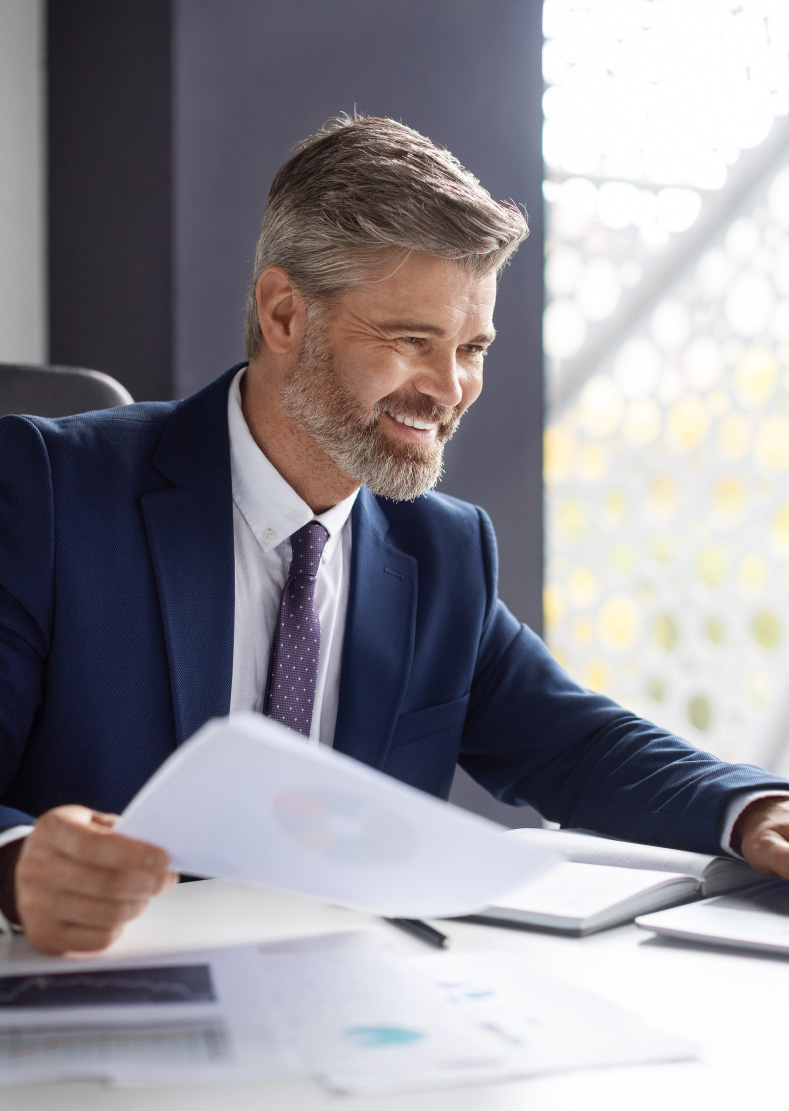 Smiling man in a navy suit at a desk reading a printed report