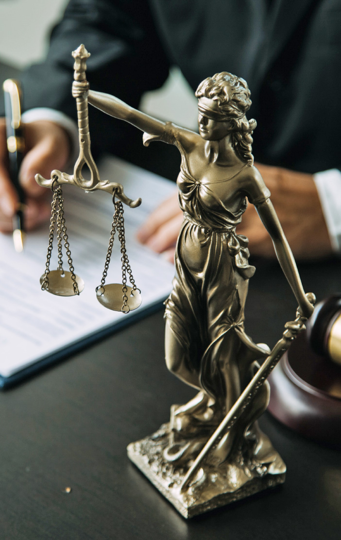 Gold Lady Justice statue holding scales beside a gavel, a person signing papers behind