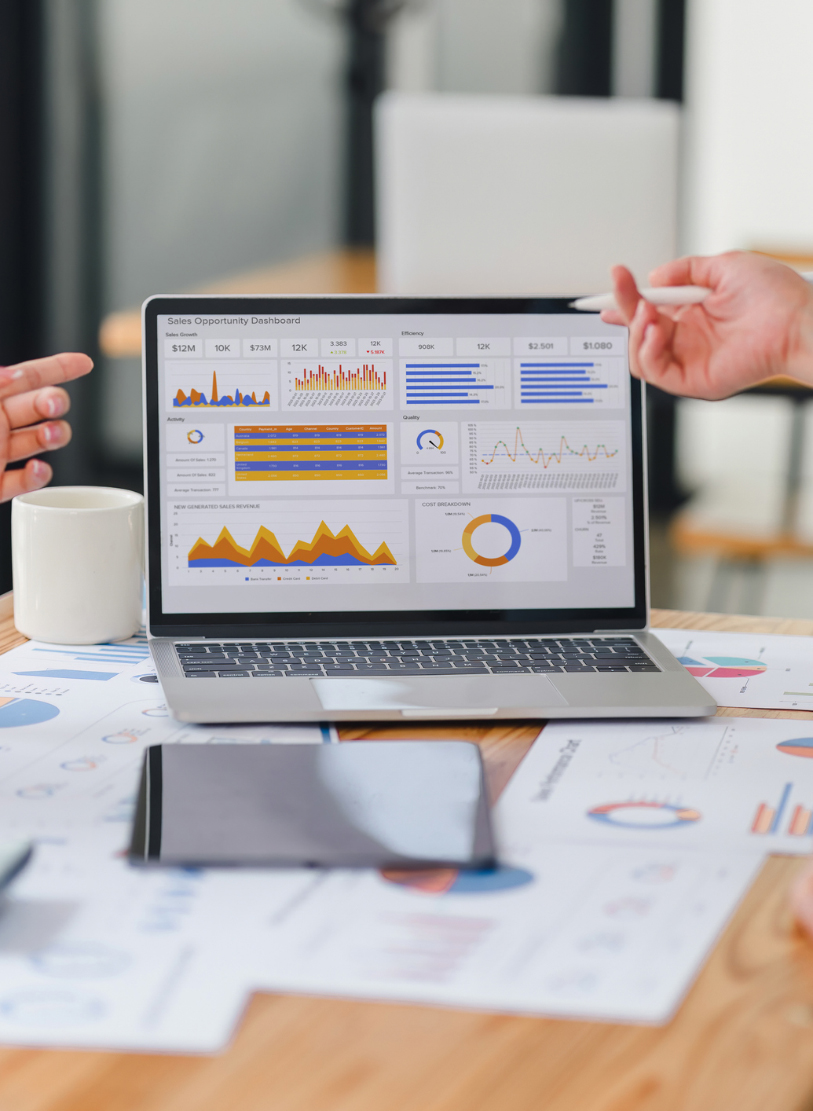 Laptop showing a Sales Opportunity Dashboard of charts and revenue figures, two people gesturing over printed reports
