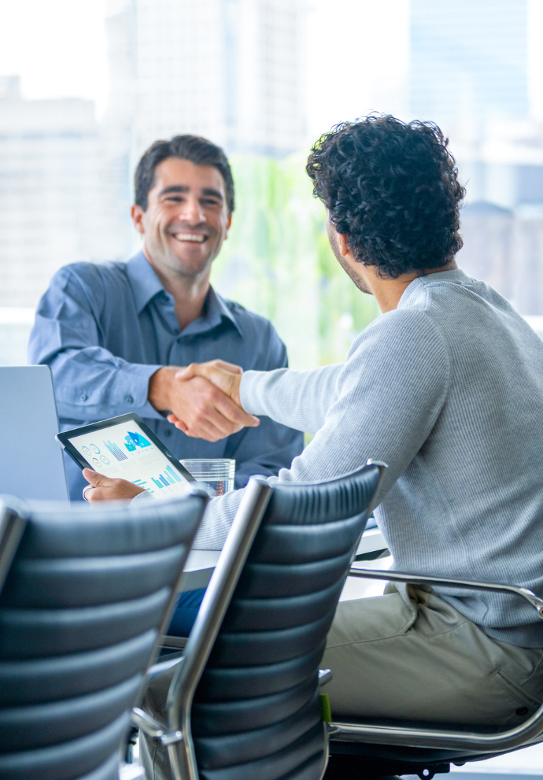 Two men shaking hands across a conference table, one holding a tablet showing charts