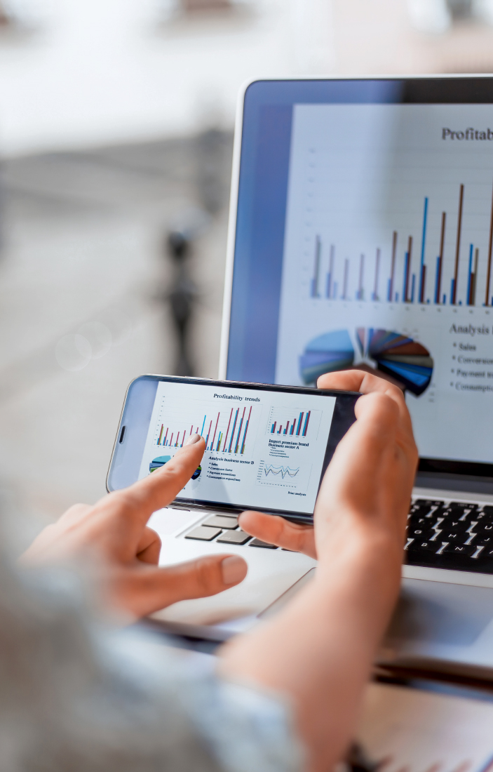 Phone showing bar charts held in front of a laptop displaying a profitability trends report