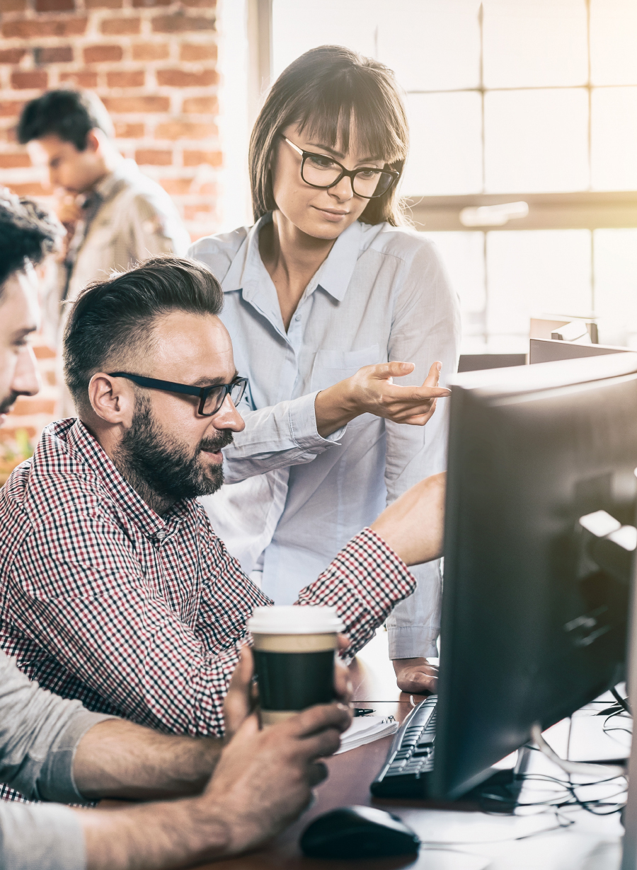 Woman pointing at a desktop monitor while a seated colleague with coffee looks on