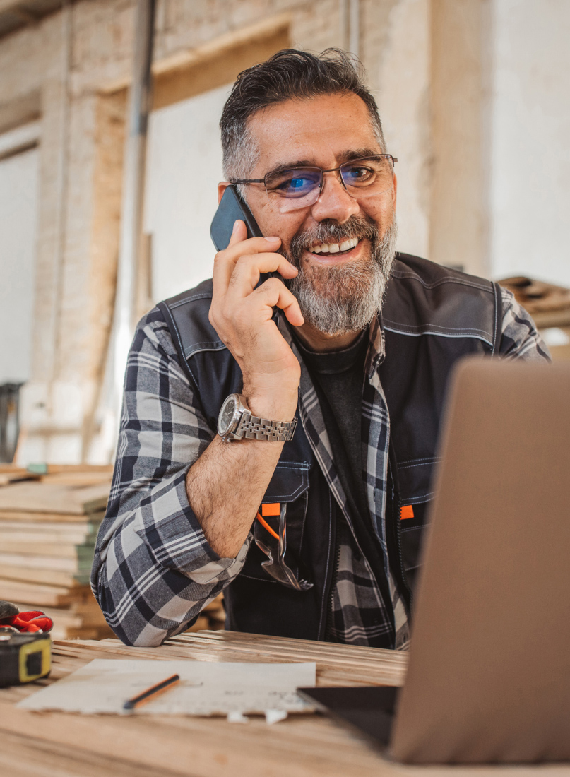 Bearded tradesman in a work vest on a phone call at a workbench, laptop and sketch in front of him
