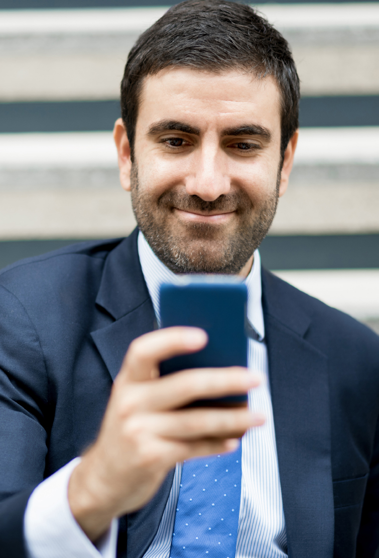 Man in a dark suit and tie smiling at his phone while sitting on outdoor steps
