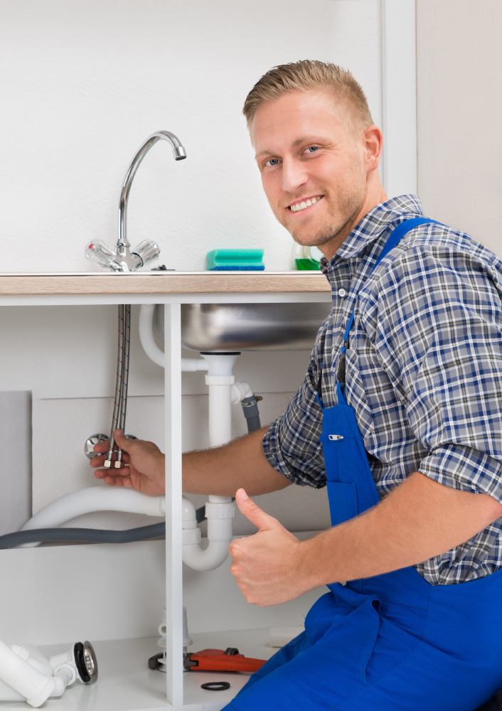 Plumber in blue overalls under a kitchen sink connecting supply hoses and giving a thumbs up