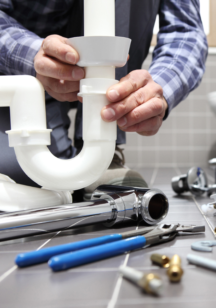 Plumber's hands fitting a white sink P-trap, pliers and fittings on the tiled floor