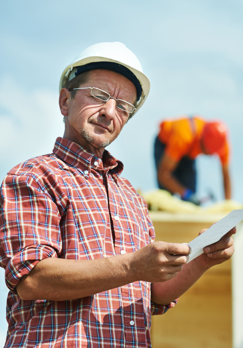 Man in a white hard hat and plaid shirt holding a paper, a roofer working behind him