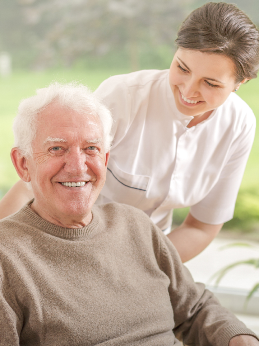 Smiling older man seated with a caregiver in white scrubs leaning over him