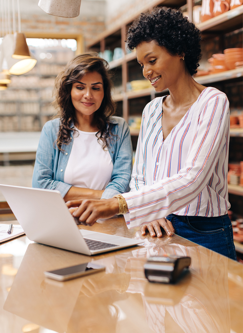 Two women behind a shop counter looking at a laptop, stocked shelves behind them