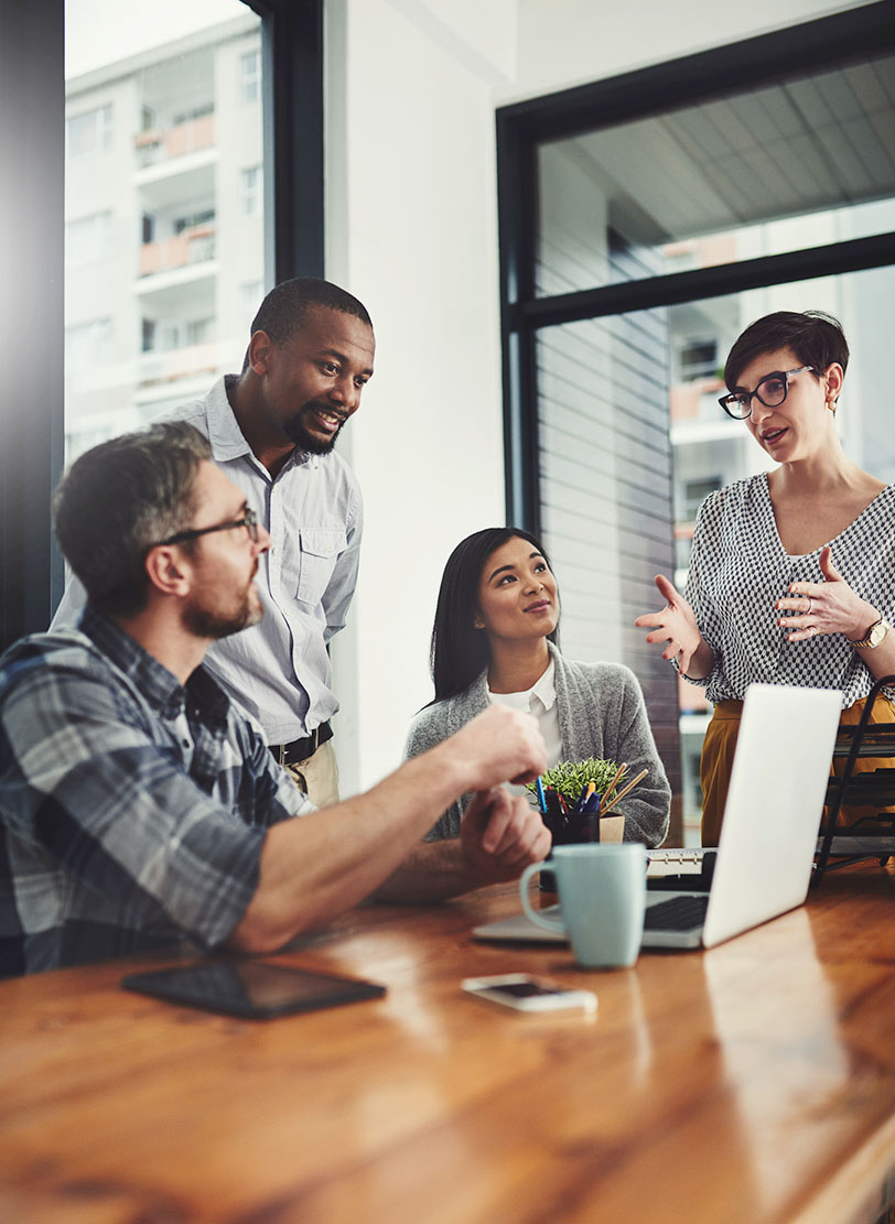 Four colleagues talking around a wooden table, one standing and gesturing as she speaks